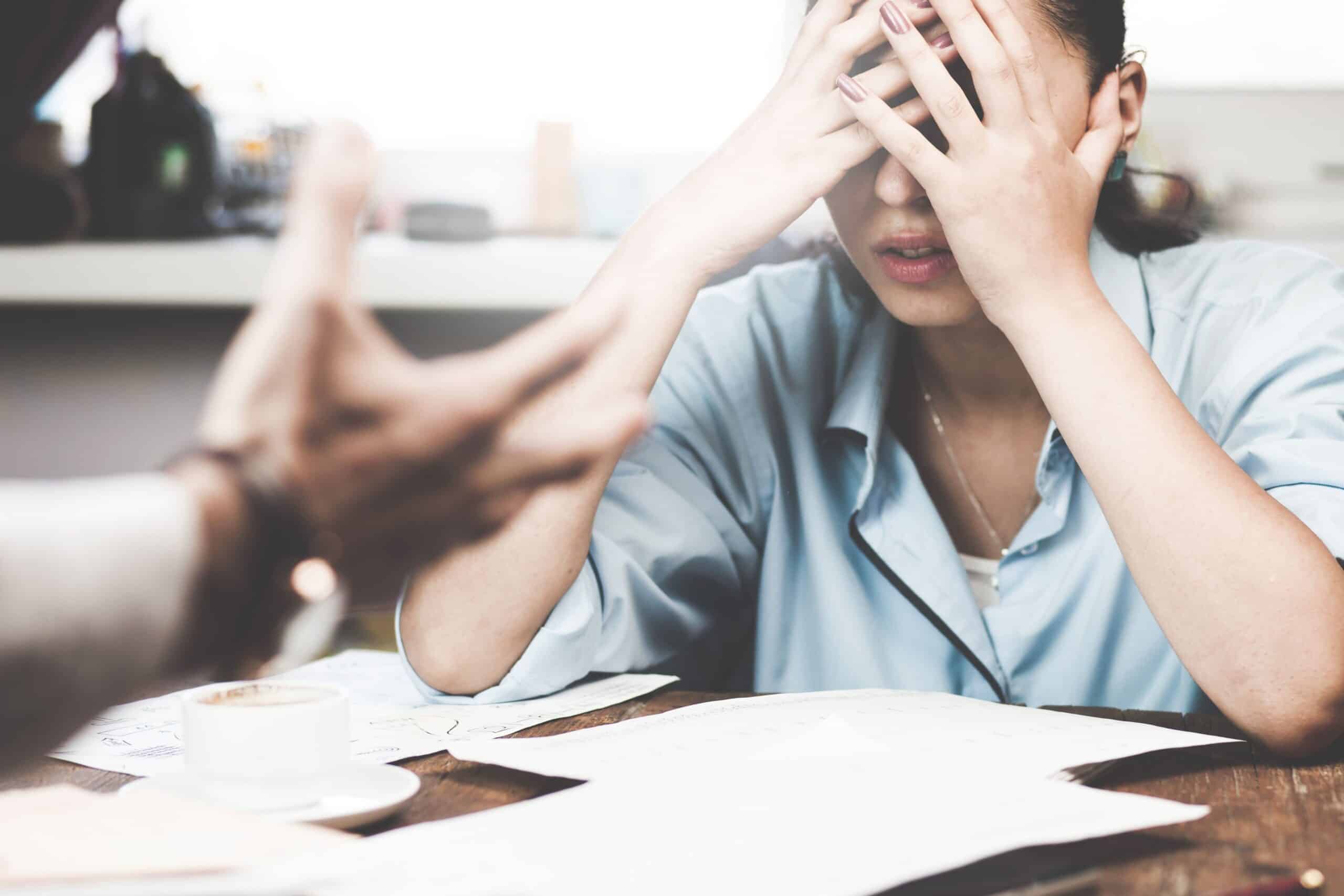 Eine Frau sitzt an einem Tisch, hält sich verzweifelt den Kopf und blickt nach unten. Im Vordergrund ist eine ausgestreckte Hand zu sehen, die auf sie gerichtet ist, was auf eine angespannte Situation oder Diskussion hinweist. **"Eine Frau sitzt an einem Tisch, hält sich verzweifelt den Kopf und blickt nach unten. Im Vordergrund ist eine ausgestreckte Hand zu sehen, die auf sie gerichtet ist, was auf eine angespannte Situation oder Diskussion hinweist. Auf dem Tisch liegen Papiere, ein Kaffee und Büromaterial."** Dieser Alt-Text beschreibt die Szene präzise für Screenreader-Nutzer und vermittelt die Emotion der Situation.