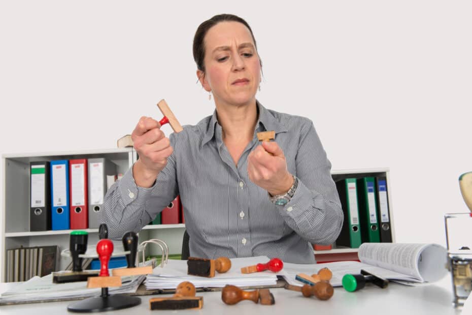 Frau in einem Büro, umgeben von Aktenordnern und Stempeln, prüft konzentriert zwei Stempel in ihren Händen.