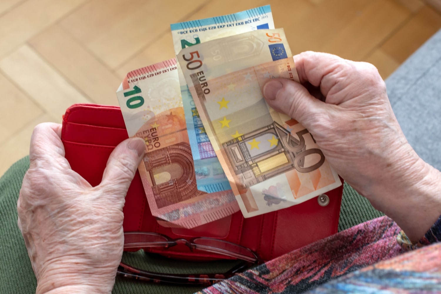 Eine ältere Person hält mehrere Euro-Geldscheine in der Hand, darunter 10-, 20- und 50-Euro-Scheine, vor einem roten Portemonnaie. Im Hintergrund ist ein Holzboden zu erkennen.