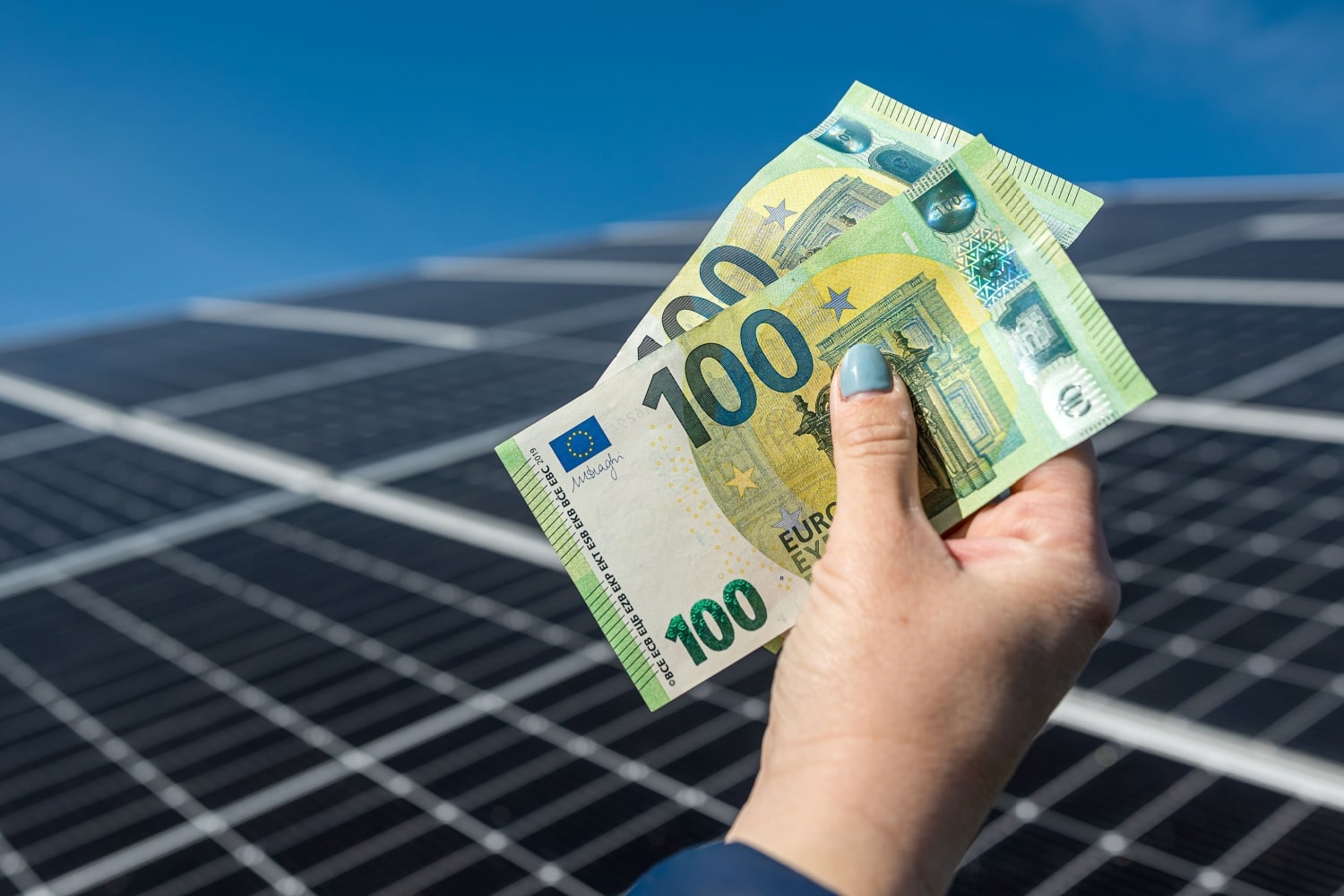 Eine Hand hält zwei 100-Euro-Scheine vor einem Hintergrund aus Solarmodulen unter klarem, blauem Himmel.