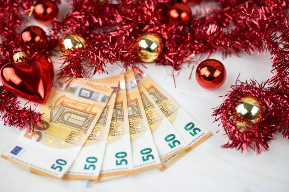 50-Euro-Scheine liegen umgeben von roter und goldener Weihnachtsdekoration, darunter Christbaumkugeln und Glitzer-Girlanden, auf einem hellen Untergrund.