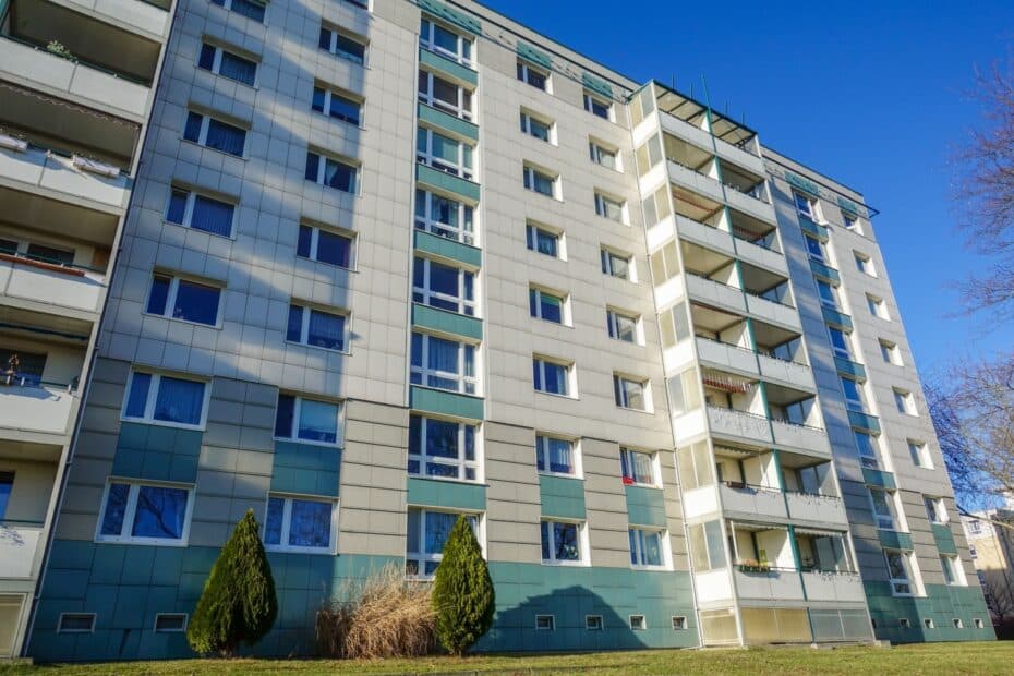 Eine mehrstöckige Wohnhausfassade mit Balkonen und Fenstern, umgeben von Grünflächen und Bäumen, bei klarem, sonnigem Wetter.